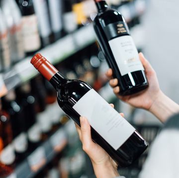over the shoulder view of woman walking through liquor aisle and choosing bottles of red wine from the shelf in a supermarket