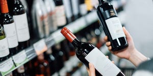 over the shoulder view of woman walking through liquor aisle and choosing bottles of red wine from the shelf in a supermarket