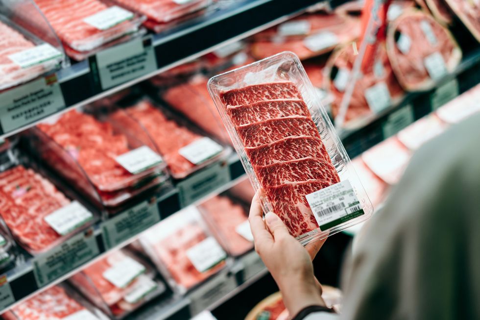 over the shoulder view of young asian woman shopping in a supermarket she is choosing meat and holding a packet of organic beef in front of the refrigerated section