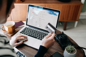 over the shoulder view of young asian woman shopping online for flight tickets on airline website with laptop, entering credit card details to make mobile payment at home camera and passport on the table travel planning booking a holiday online