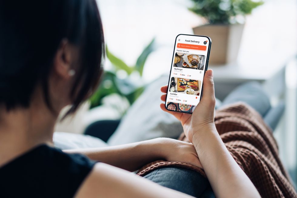 over the shoulder view of young asian woman sitting on the sofa, using meal delivery service and ordering food online with smartphone mobile app at home