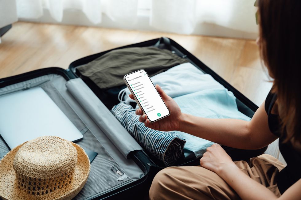 over the shoulder view of young woman looking at checklist on smartphone while packing luggage