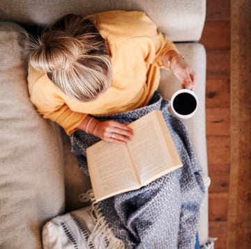 Overhead Shot Looking Down On Woman At Home Lying On Reading Book And Drinking Coffee