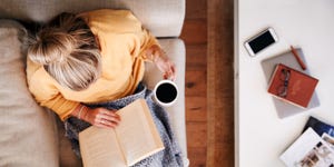 Overhead Shot Looking Down On Woman At Home Lying On Reading Book And Drinking Coffee