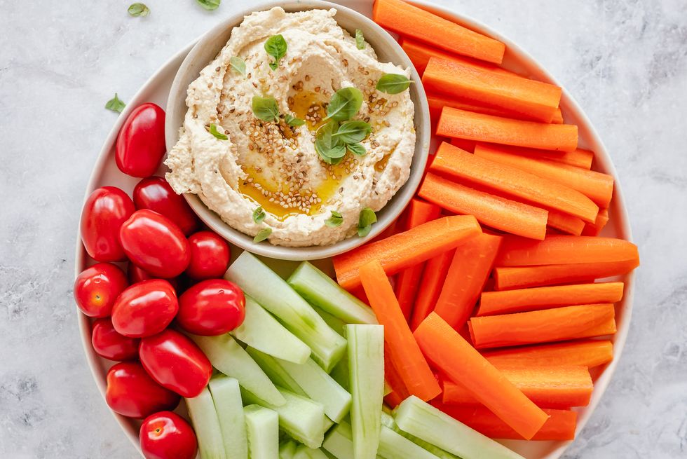 overhead view of a bowl of hummus with cherry tomatoes, carrot and cucumber batons