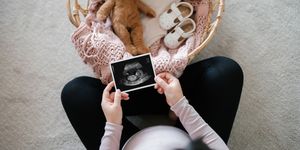 overhead view of pregnant woman holding an ultrasound scan photo of her baby, with a moses basket filling with baby clothing, baby gift