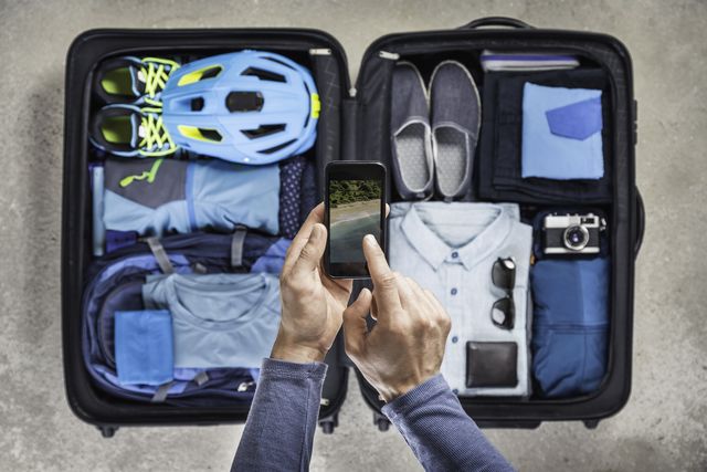 Overhead view of mans hands using smartphone touchscreen above packed suitcase with blue bike helmet, backpack, retro camera and shirt