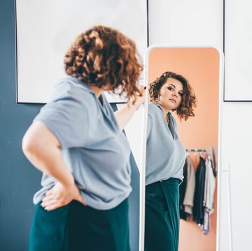 overweight young woman in glasses admires choice of clothes standing in front of large mirror in stylish room reflection view