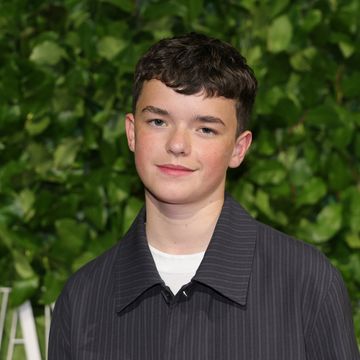 a young boy smiles at the camera while standing in front of a leafy green wall, he wears a black striped collared shirt over a white tshirt