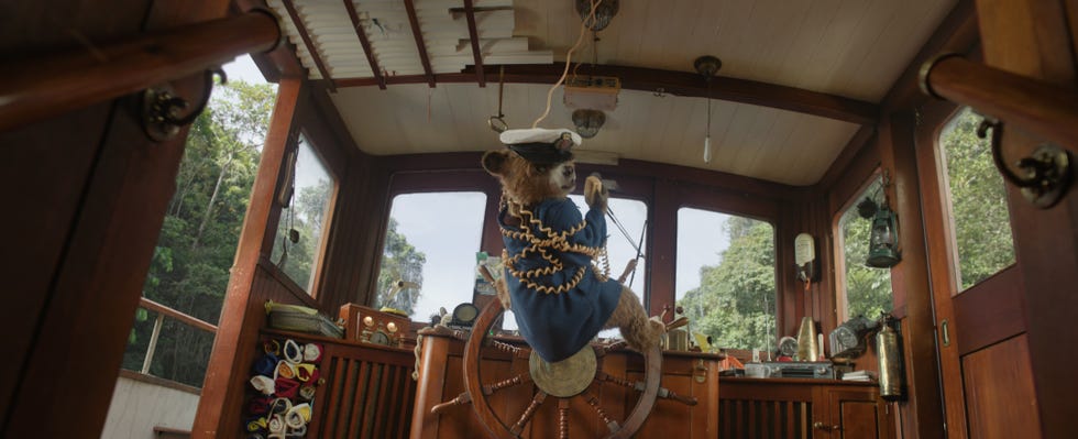 paddington bear wearing a captains hat and a blue coat is steering a boat, paddington in peru