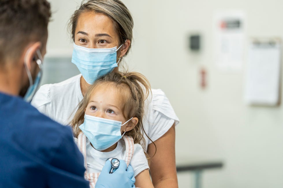 paediatrician doctor examining a young female patient sitting on her mothers lap wearing masks