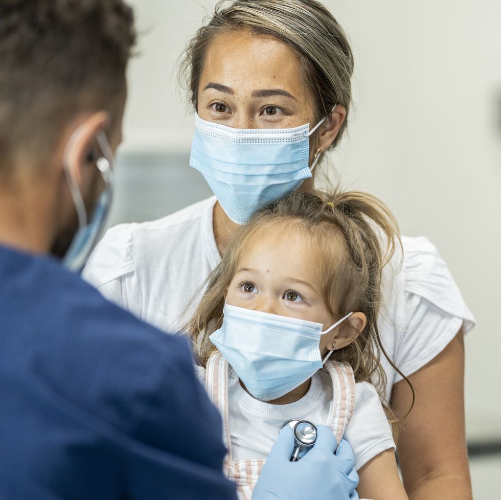 paediatrician doctor examining a young female patient sitting on her mothers lap wearing masks