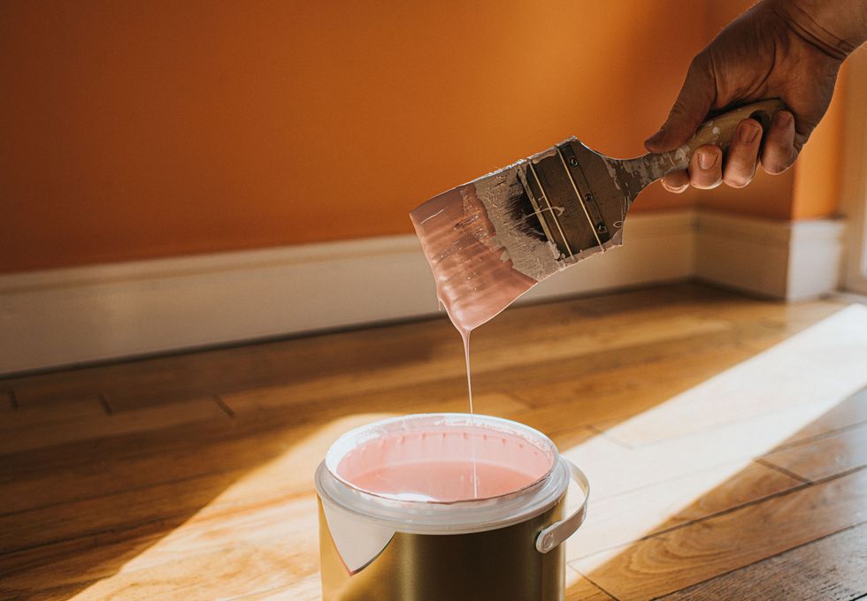 hand holds a thick paint brush and removes it, after dipping it in the paint it creates a droplet of paint which splashes back into the pot the wall behind is painted orange and the colour in the tin is pink, which clashes conceptual with space for copy
