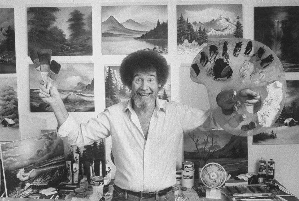 a smiling bob ross stands in a studio with his arms raised, he is holding three paint brushes in one hand and a palette with paint blots in the other, behind him is a desk full of supplies and a wall with his paintings