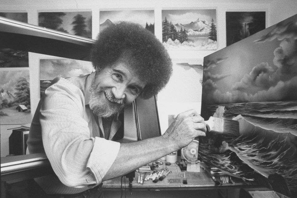 bob ross in a painting studio, smiling into the camera, holding a paint brush against a painting of an ocean landscape