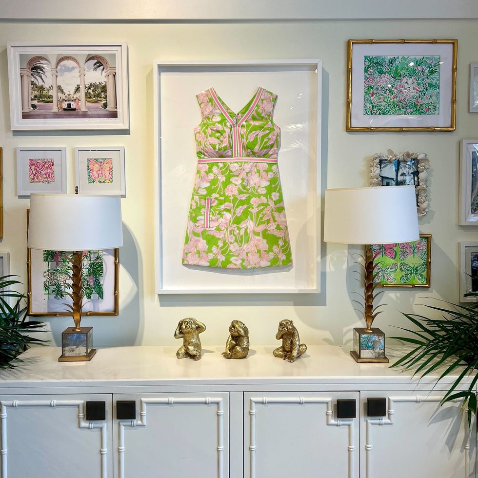 a white kitchen dresser with lamps on it next to a wall with a framed dress and framed colorful prints