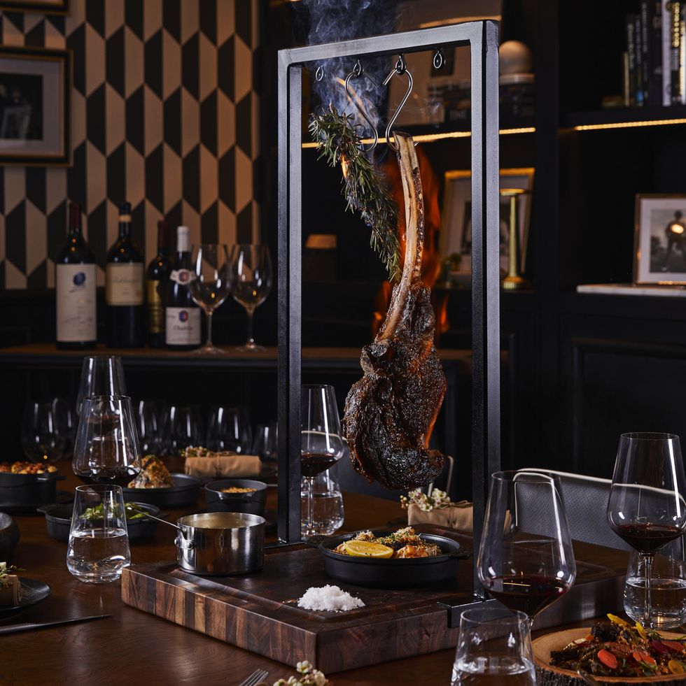 a tomahawk steak hanging from a device over a table with wine glasses and side dishes on it
