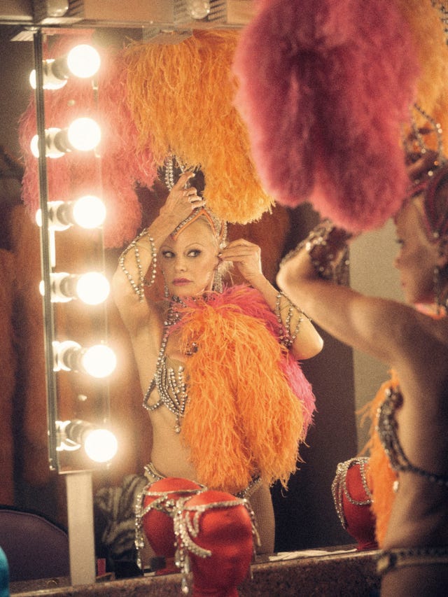 a performer preparing in front of a vanity mirror adorned with bright feathers