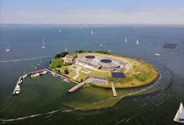 het nieuwe bezoekers centrum afgebeeld tussen de twee aanlegsteigers zal worden verzonken in de wal van pampus waardoor bezoekers vanaf het ijmeer weer vrij zicht hebben op de historische geschutskoepels