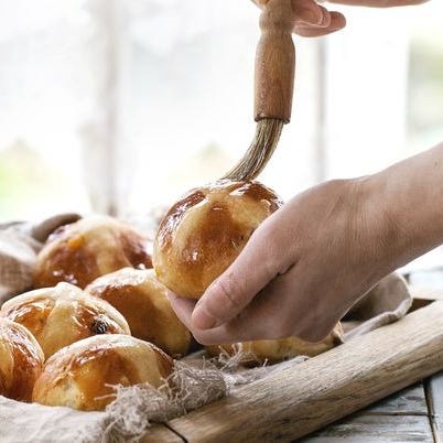 homemade easter traditional hot cross buns on wooden tray with textile over white wooden table window at background female hands cover with syrup natural day light rustic style