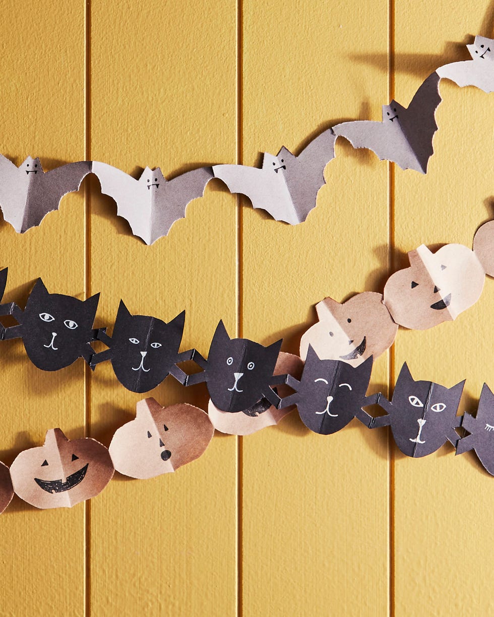 papers chains of bats cut from gray paper cats cut from black paper and pumpkins cut from kraft paper hanging on a yellow slated wall