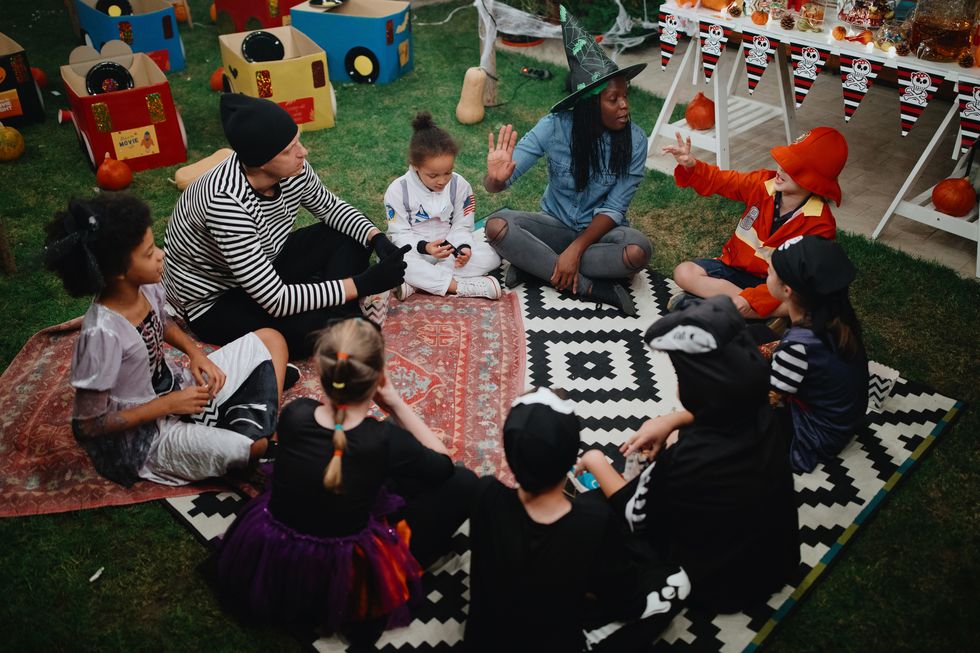 parents telling scary stories to kids at outdoor halloween party they are sitting cross legged on rugs on the grass, one parent is in a witch's hat