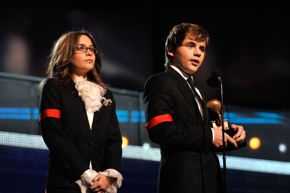 paris jackson stands with her hands clasp on a stage, prince jackson speaks into a microphone to the right while holding a grammy award, both children wear black suits with a red arm band on one sleeve and white dress shirts