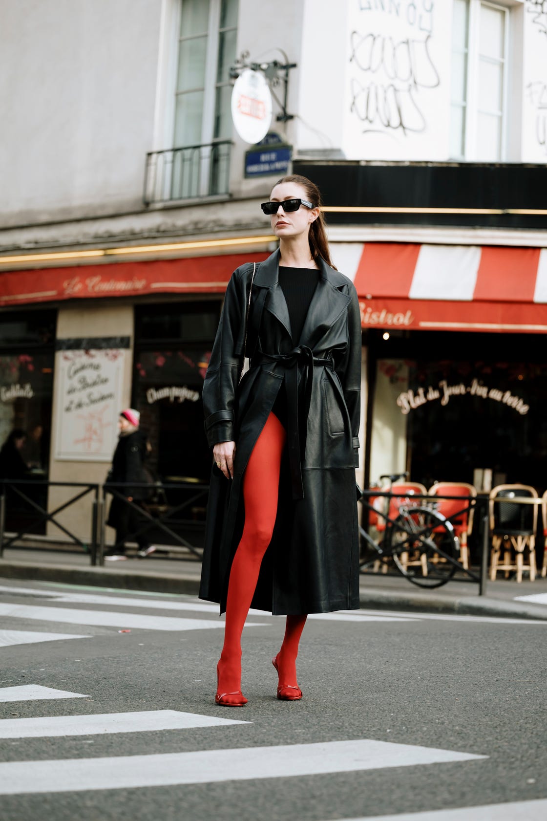 a figure in a stylish black leather trench coat and red leggings stands at a crosswalk