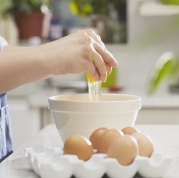 partial view of woman wearing an apron in a kitchen, cracking an egg into a bowl