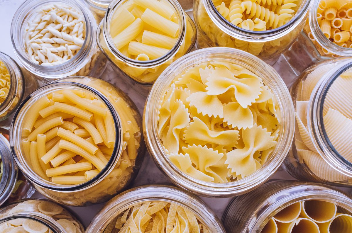 various types of pasta, including tube shapes, large shells, and bow tie shapes, in glass jars on marble background
