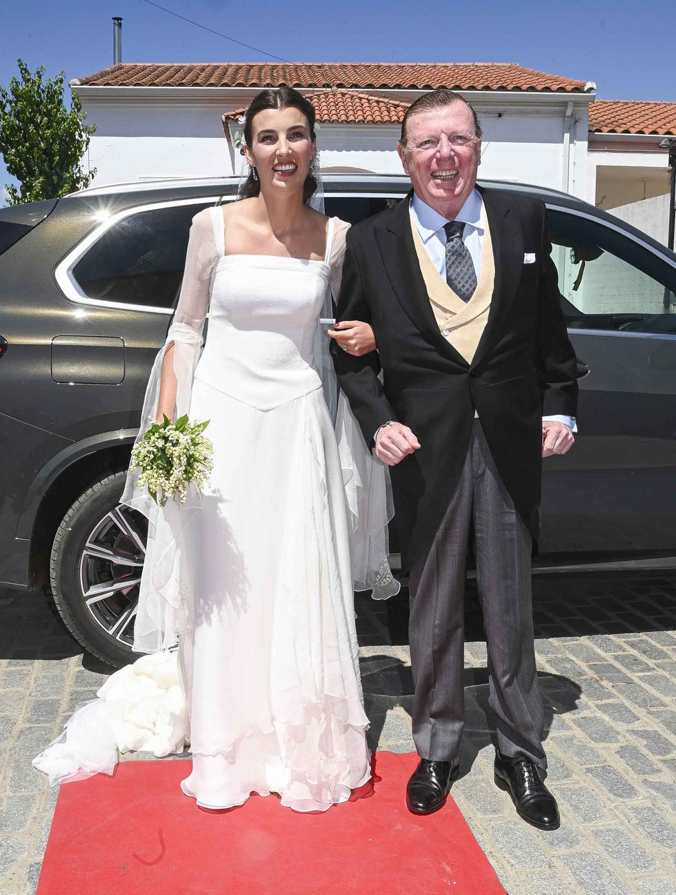 couple dressed in formal attire standing on a red carpet outside a vehicle