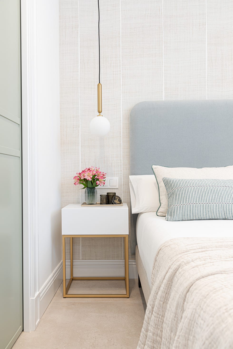 cama con cabecero azul, papel pintado tejido a rayas, mesita de noche blanca con estructura de metal dorado y lámpara suspendida de bola