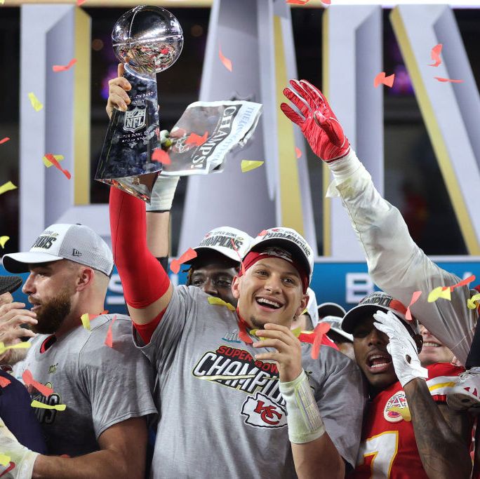 patrick mahomes lifts a silver football trophy in the air and smiles, he wears a gray tshirt and white ballcap, several people surround him as confetti rains down