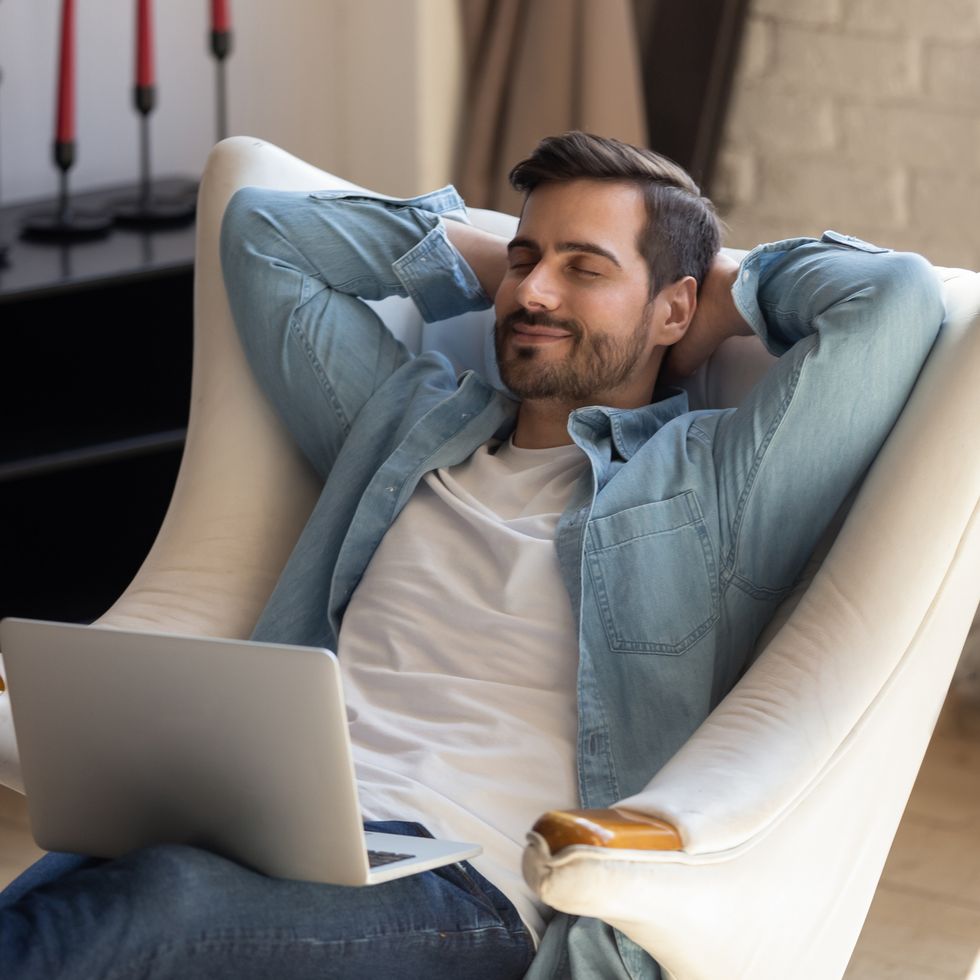 peaceful young man daydreaming in comfortable armchair, holding computer