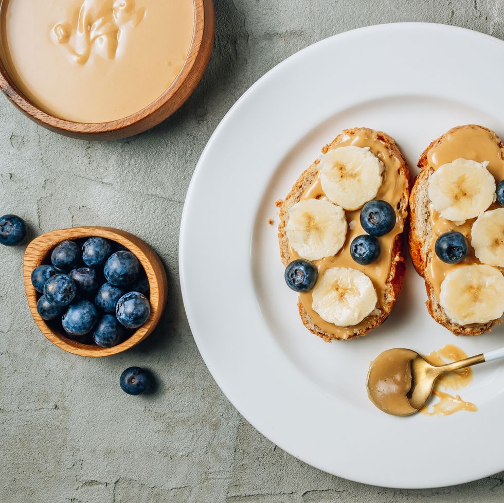 buckwheat healthy bread with peanut butter, banana and blueberry on white plate over concrete background top view flat lay summer breakfast