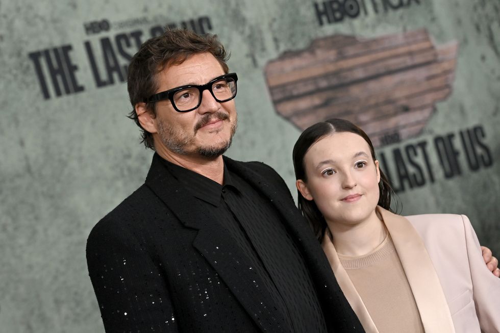 pedro pascal, wearing a black suit, black shit, and black rimmed glasses, stands with his arm around bella ramsey, who wears a peach suit jacket and shirt
