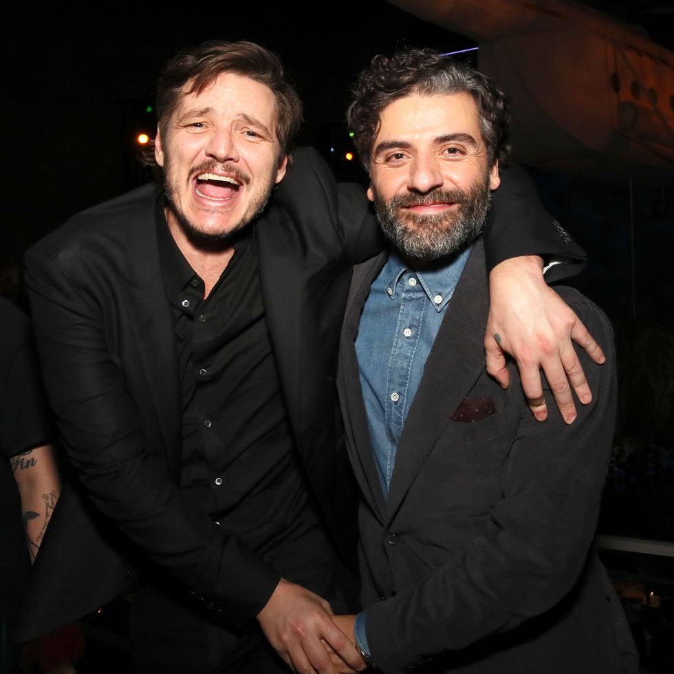 pedro pascal, wearing a black suit jacket and shirt, laughs and puts his arm around oscar isaac, who wears a black suit jacket and denim blue shirt
