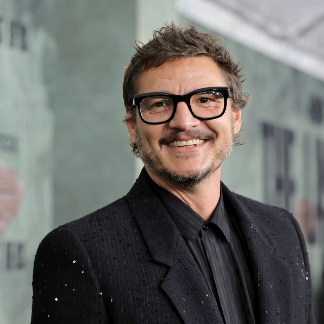 pedro pascal turns his head toward the camera and smiles, he wears black thick framed glasses and a black suit jacket with sequins over a black and dark gray collared shirt