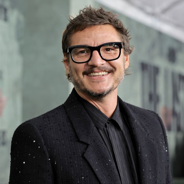 pedro pascal turns his head toward the camera and smiles, he wears black thick framed glasses and a black suit jacket with sequins over a black and dark gray collared shirt