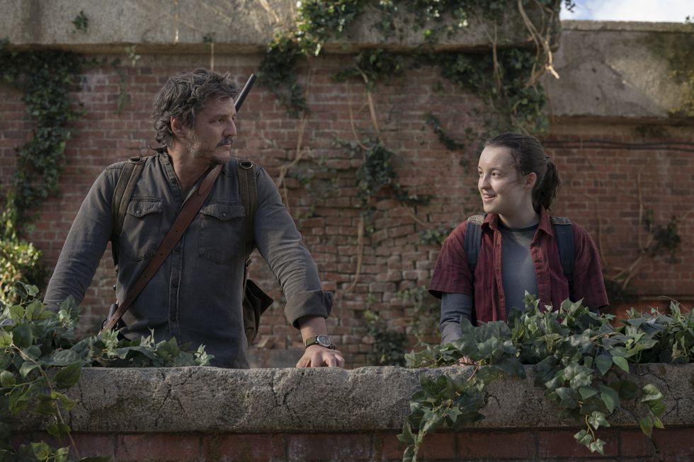 pedro pascal and bella ramsey in character for the last of us, they stand in front of a brick way and look at each other