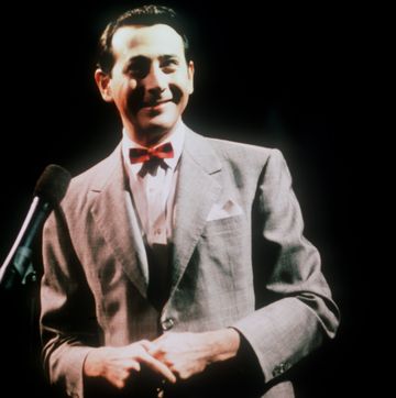 paul reubens as pee wee herman on a darkened stage, standing in front of a microphone and smiling