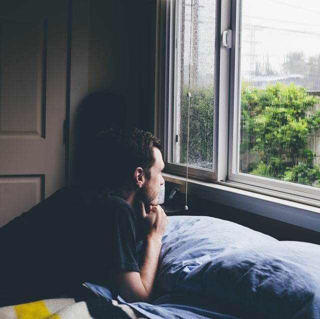 man lying on bed staring through window at rain