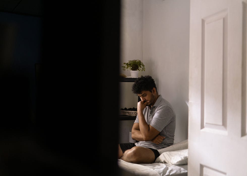 pensive and anxious hispanic man sitting on a bed in a room