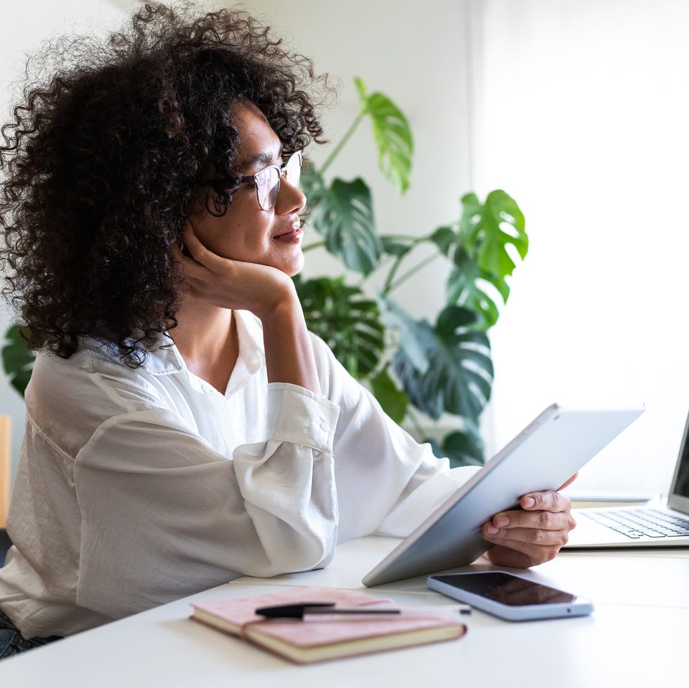 pensive, happy multiracial young woman working at home office using multiple devices: digital tablet, laptop and phone.