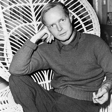 truman capote sits in a highbacked wicked chair and looks at the camera as he rests his head on one hand, he wears a sweater over a collared shirt and slacks