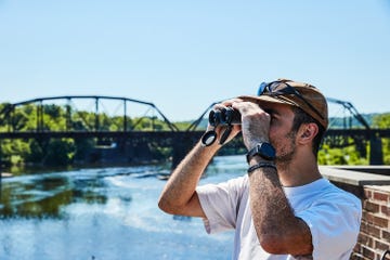 A person using binoculars to observe a scene by the river near a bridge