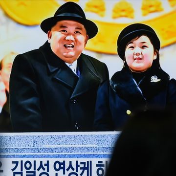 several people watch a television screen, upon which kim jong un and his daughter kim ju ae stand at a podium and look upwards, both wearing black coats and hats