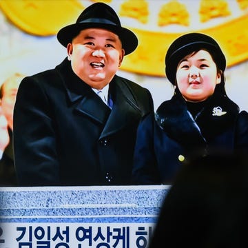 A news broadcast featuring Kim Jong-un and Kim Ju-Ae several people watch a television screen, upon which kim jong un and his daughter kim ju ae stand at a podium and look upwards, both wearing black coats and hats
