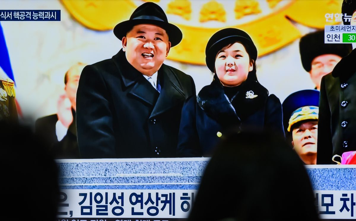 several people watch a television screen, upon which kim jong un and his daughter kim ju ae stand at a podium and look upwards, both wearing black coats and hats