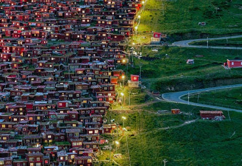 Deze foto werd genomen in de boeddhistische academie van Seda Larung Gar Vanuit de dichtstbijzijnde stad is het ongeveer veertien uur rijden naar Larung Gar en de reis over bergwegen is zwaar In deze opname zijn aan de linkerzijde de rode huisjes te zien terwijl aan de andere zijde een lege weg zich door het groene landschap slingert Op hun terugkeer naar huis volgen de monniken de lampen Ik had het geluk om dit gebied vast te leggen en was diep geraakt door het geloof van de monniken Ik ben van plan volgende zomer naar Seda terug te keren om meer fotos te maken